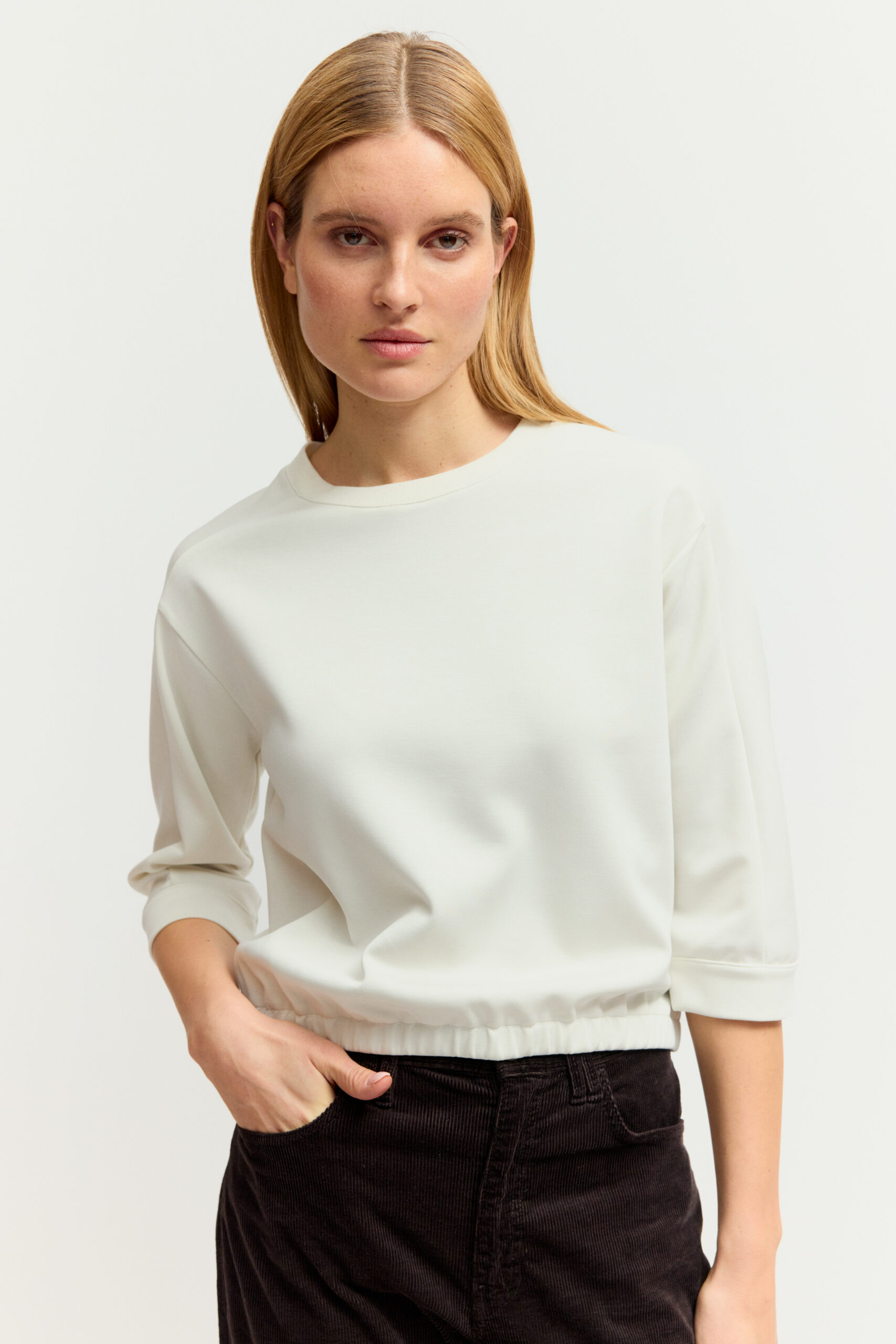 Eine junge Frau mit blonden Haaren trägt ein cremeweißes Sweatshirt mit dreiviertelangen Ärmeln und Bund unten sowie eine dunkle Hose vor hellem Hintergrund.
