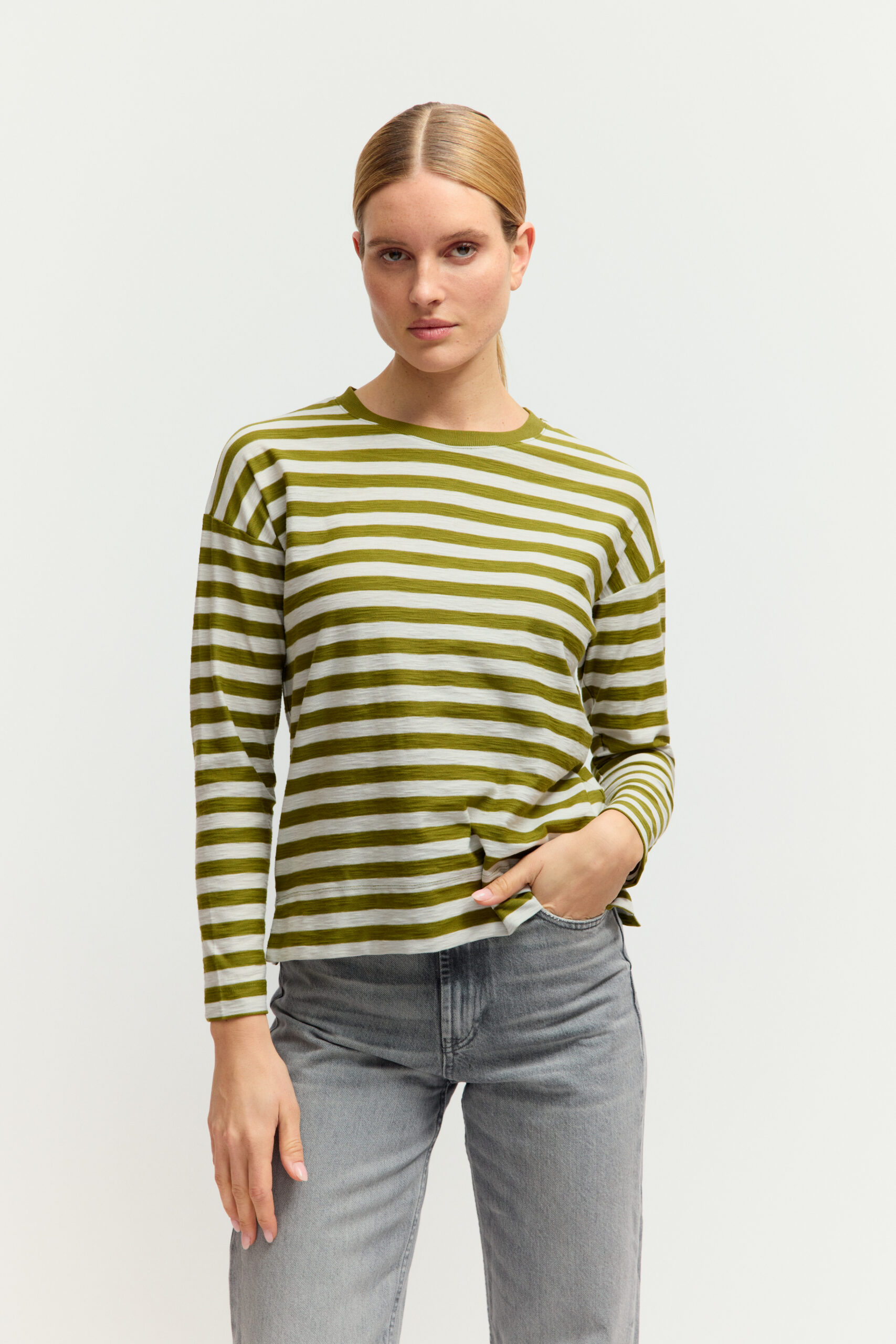 Nahaufnahme einer jungen Frau mit blondem Zopf, die ein langärmligen Feinstrickpullover mit olivgrün-weißem Querstreifenmuster vor hellem Hintergrund trägt.