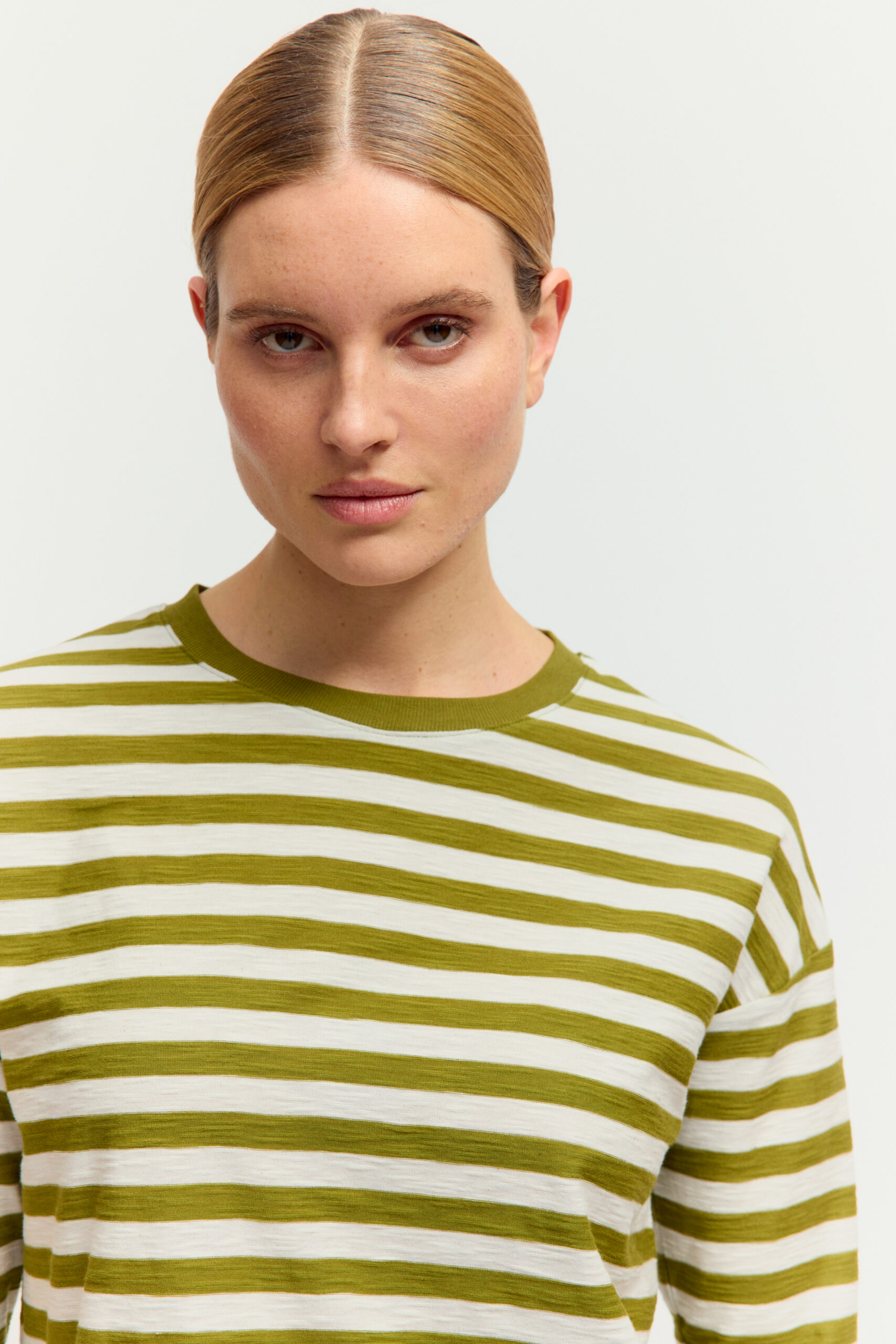 Nahaufnahme einer jungen Frau mit blondem Zopf, die ein langärmligen Feinstrickpullover mit olivgrün-weißem Querstreifenmuster vor hellem Hintergrund trägt.