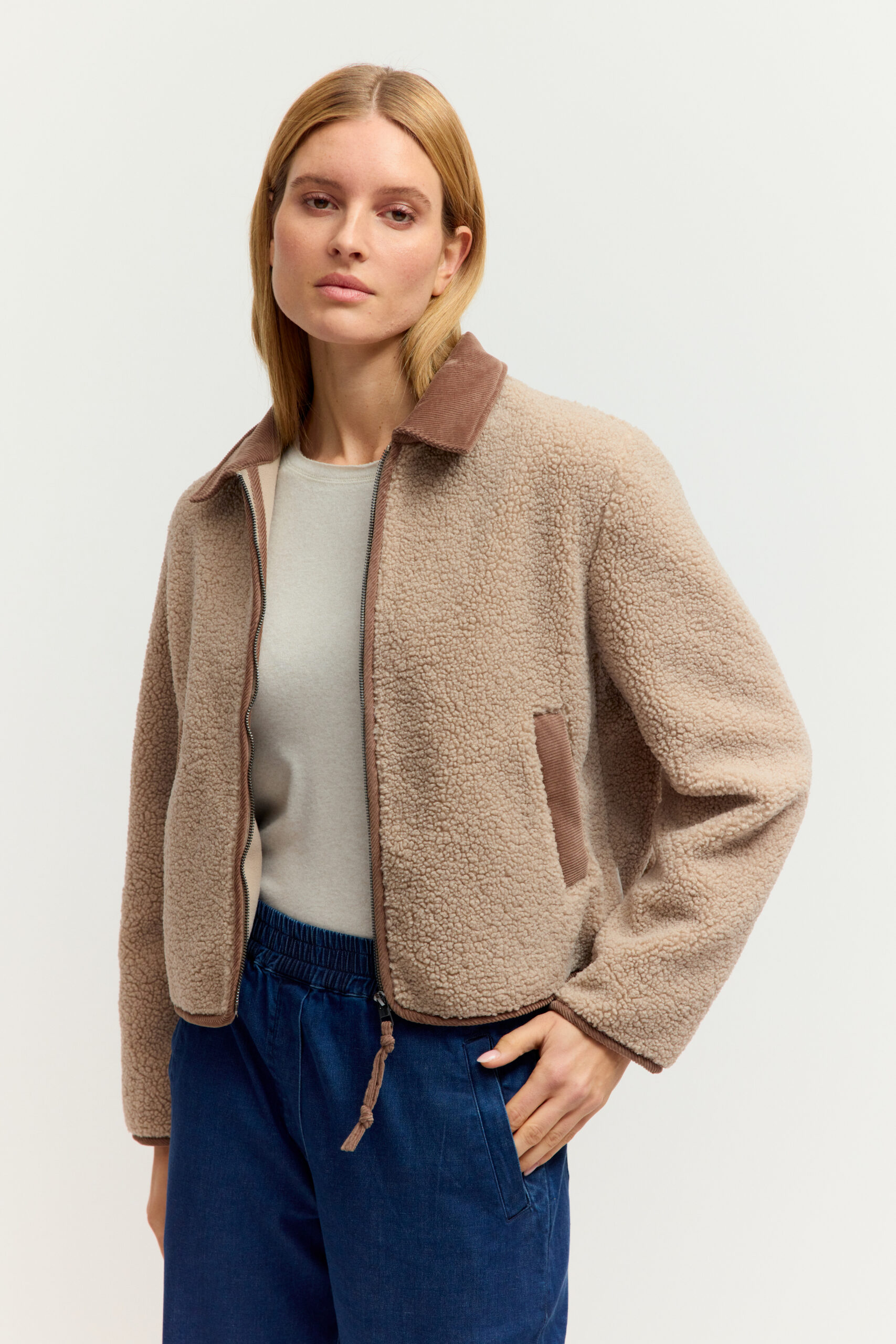 Eine junge Frau mit blonden Haaren trägt eine flauschige, beige Teddyfleece-Jacke mit braunem Kragen und Reißverschluss über einem hellen Oberteil vor hellem Hintergrund.