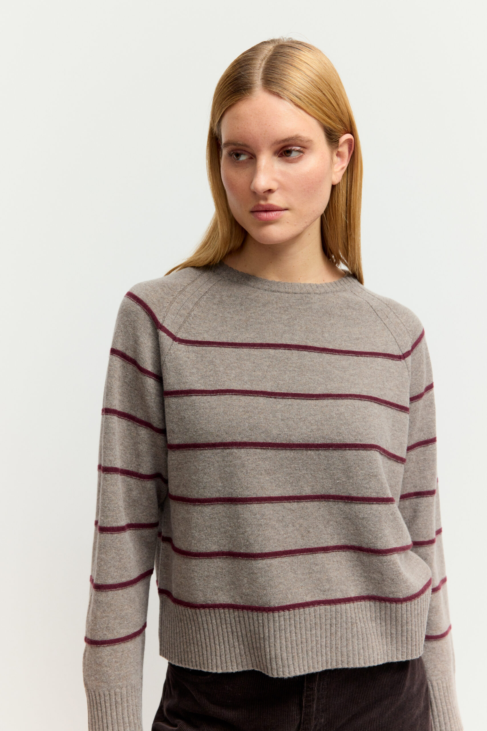 Eine junge Frau mit blonden Haaren trägt einen grau- beigen Strickpullover mit breiten, dunkelroten Querstreifen und rundem Halsausschnitt vor hellem Hintergrund.