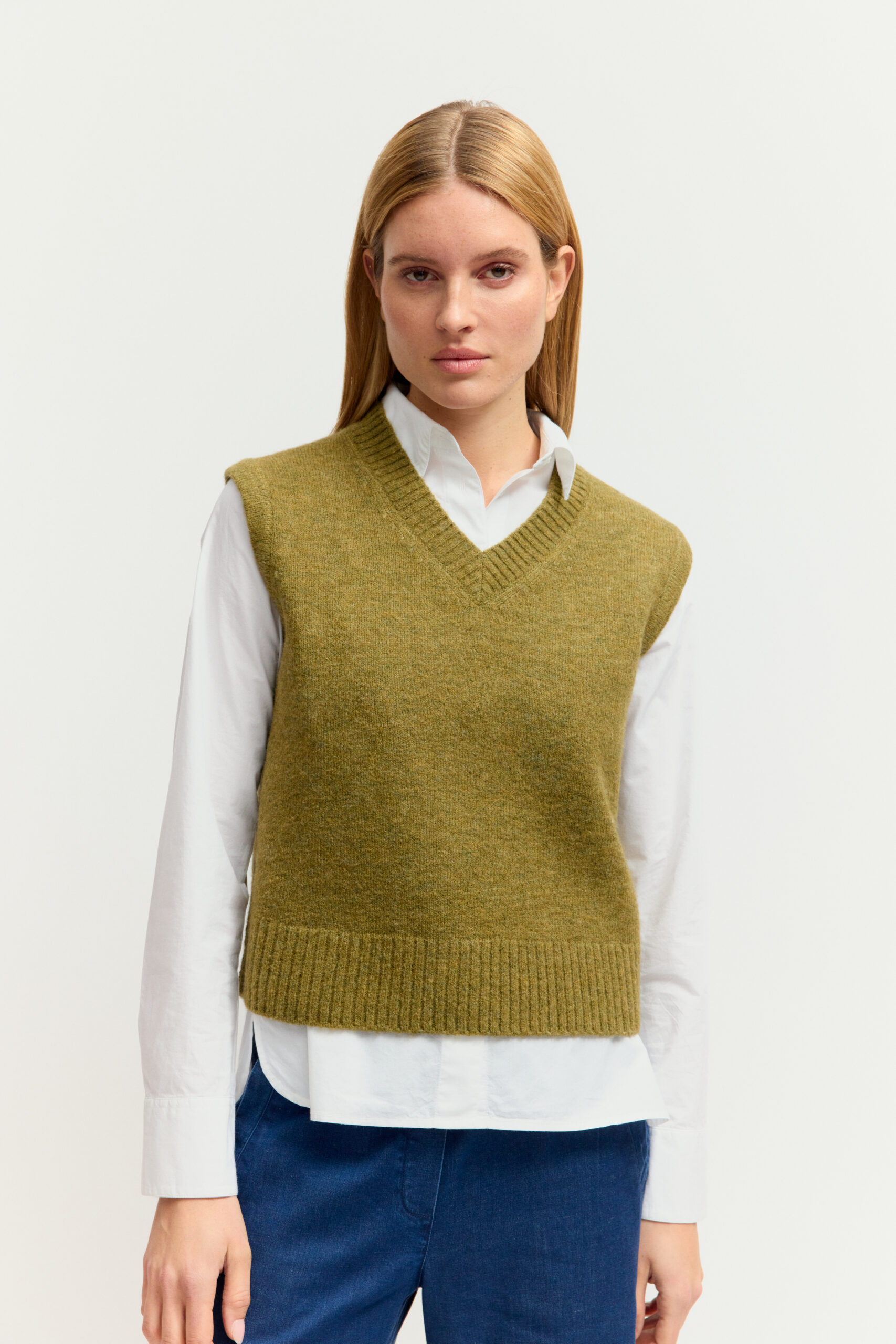 Eine junge Frau mit langen blonden Haaren präsentiert ein Outfit aus weißer Bluse, olivgrünem Strickpullover-Ärmellos (Weste) mit V-Ausschnitt, dunkler Jeans und Sneakers vor hellem Hintergrund.