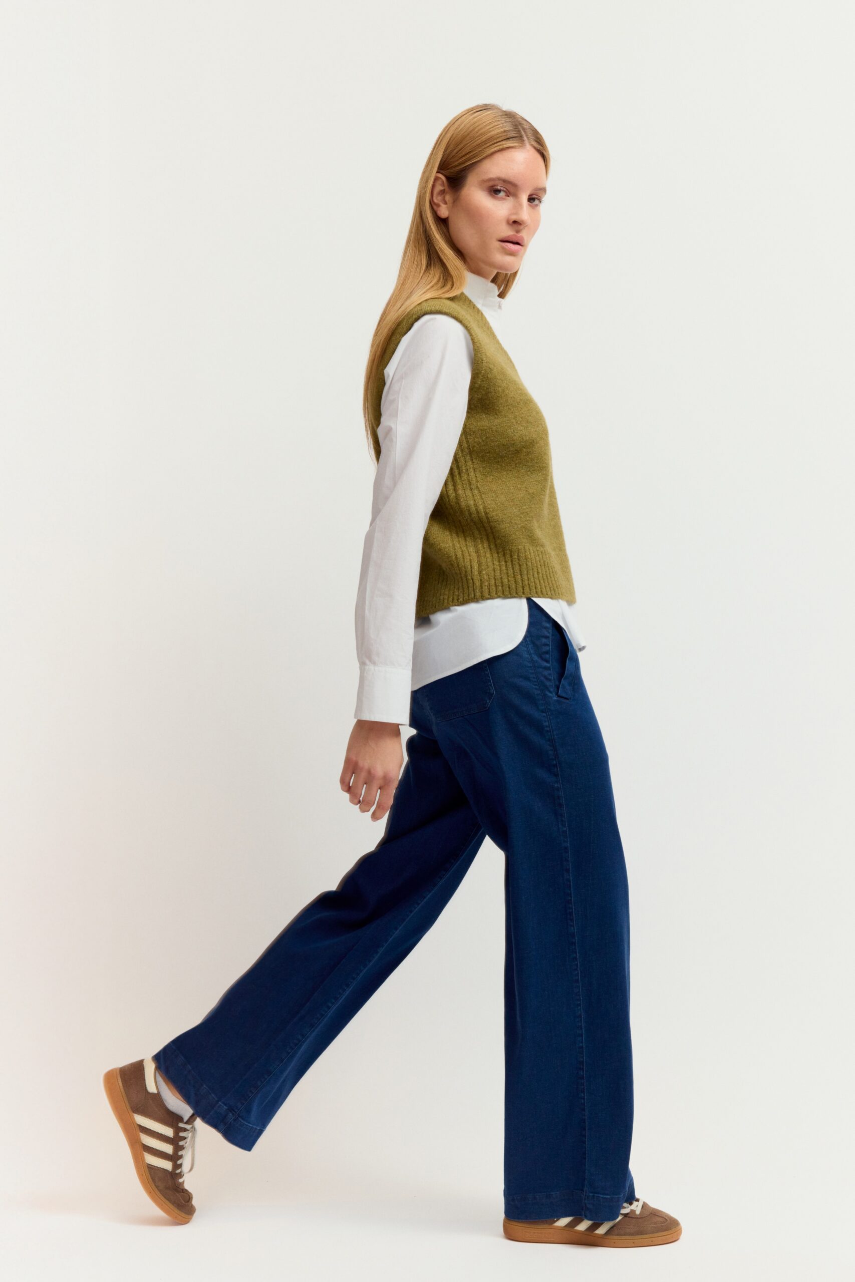 Eine junge Frau mit langen blonden Haaren präsentiert ein Outfit aus weißer Bluse, olivgrünem Strickpullover-Ärmellos (Weste) mit V-Ausschnitt, dunkler Jeans und Sneakers vor hellem Hintergrund.