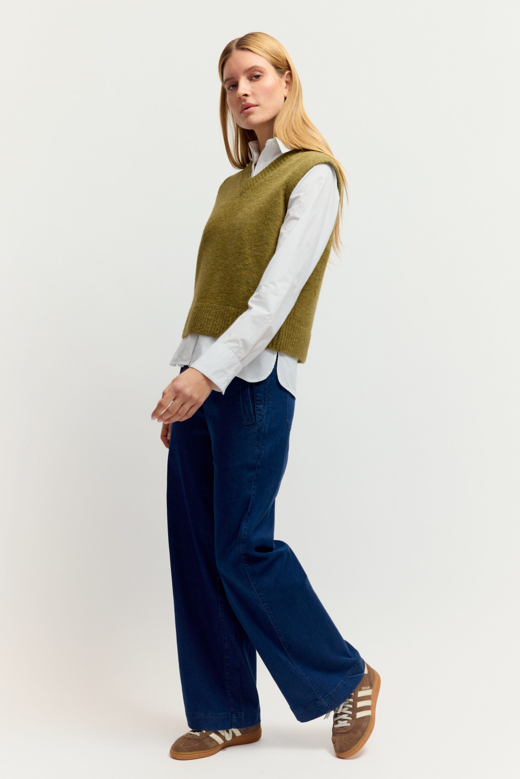 Eine junge Frau mit langen blonden Haaren präsentiert ein Outfit aus weißer Bluse, olivgrünem Strickpullover-Ärmellos (Weste) mit V-Ausschnitt, dunkler Jeans und Sneakers vor hellem Hintergrund.