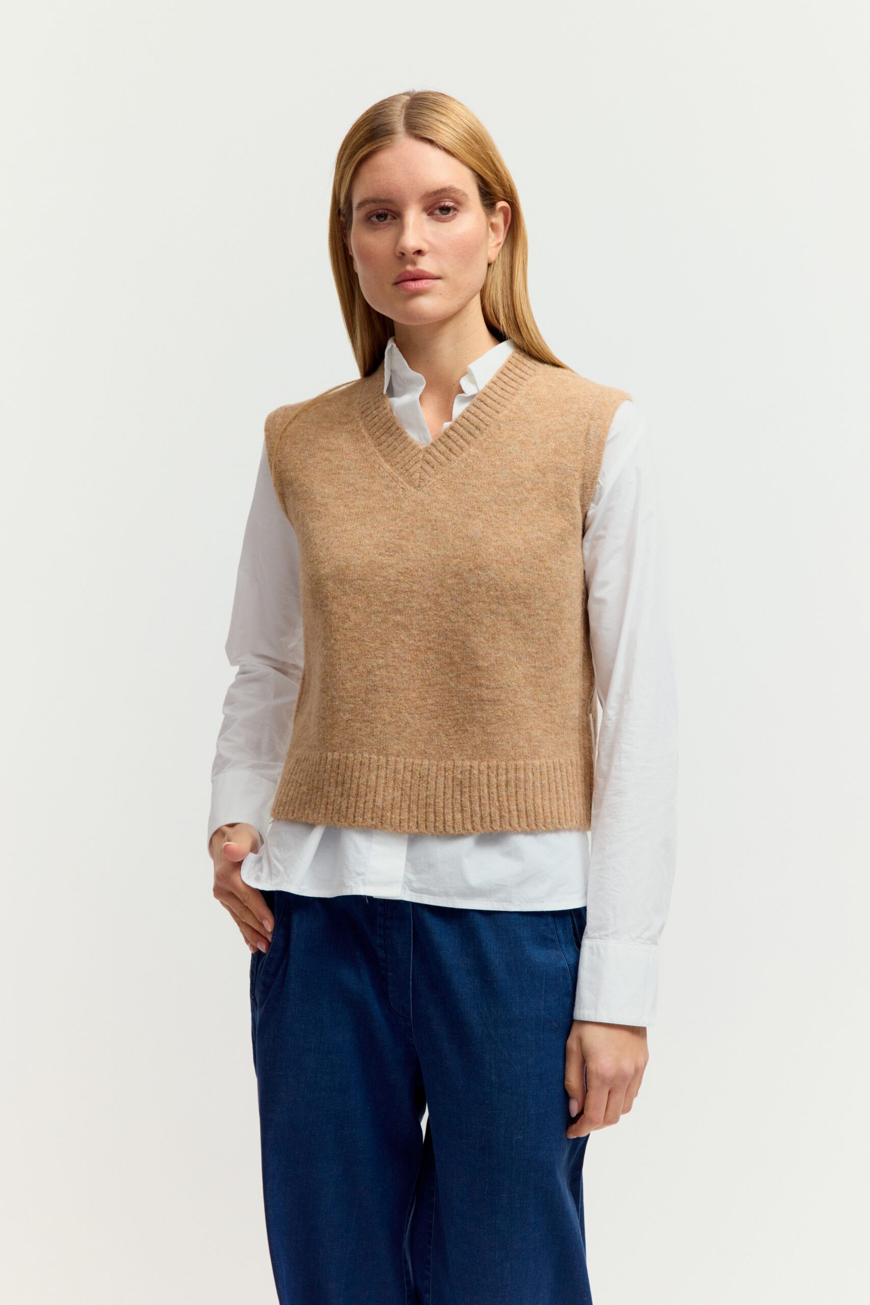 Eine junge Frau mit langen blonden Haaren präsentiert ein Outfit aus weißer Bluse, beigen Strickpullover-Ärmellos (Weste) mit V-Ausschnitt, dunkler Jeans und Sneakers vor hellem Hintergrund.