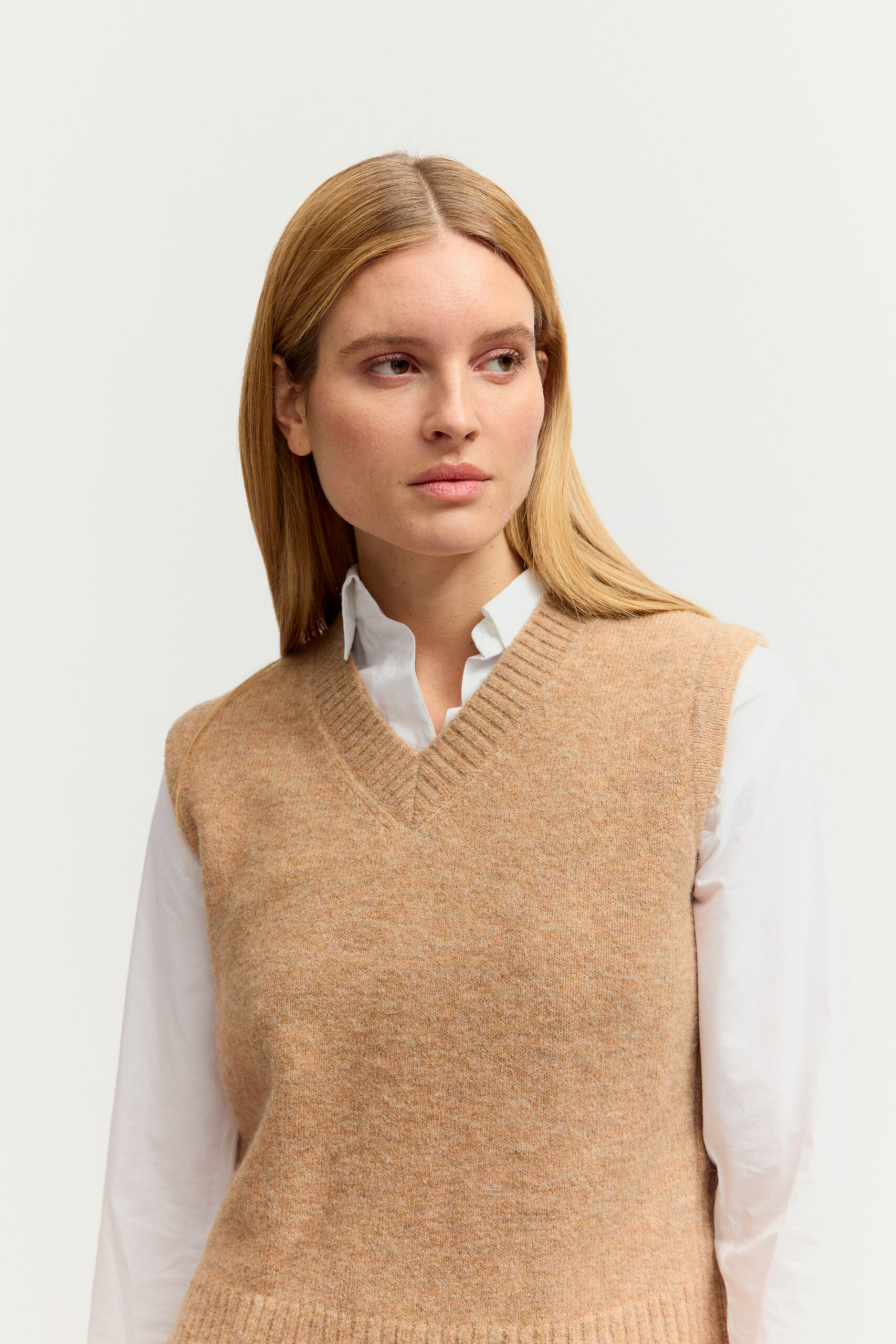 Eine junge Frau mit langen blonden Haaren präsentiert ein Outfit aus weißer Bluse, beigen Strickpullover-Ärmellos (Weste) mit V-Ausschnitt, dunkler Jeans und Sneakers vor hellem Hintergrund.